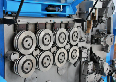 Imagem de uma máquina industrial destinada ao fabrico de molas de compressão em arame grosso, equipada com vários rolos e mecanismos de alimentação e conformação do arame metálico.